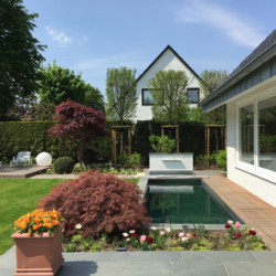 Wasserbecken Sonderanfertigung Ein moderner Hinterhof mit einem rechteckigen Pool, einem Holzdeck, einem angelegten Garten und einer Terrasse mit Stühlen. Der Bereich ist umgeben von grünem Gras, Bäumen und einem weißen zweistöckigen Haus im Hintergrund.